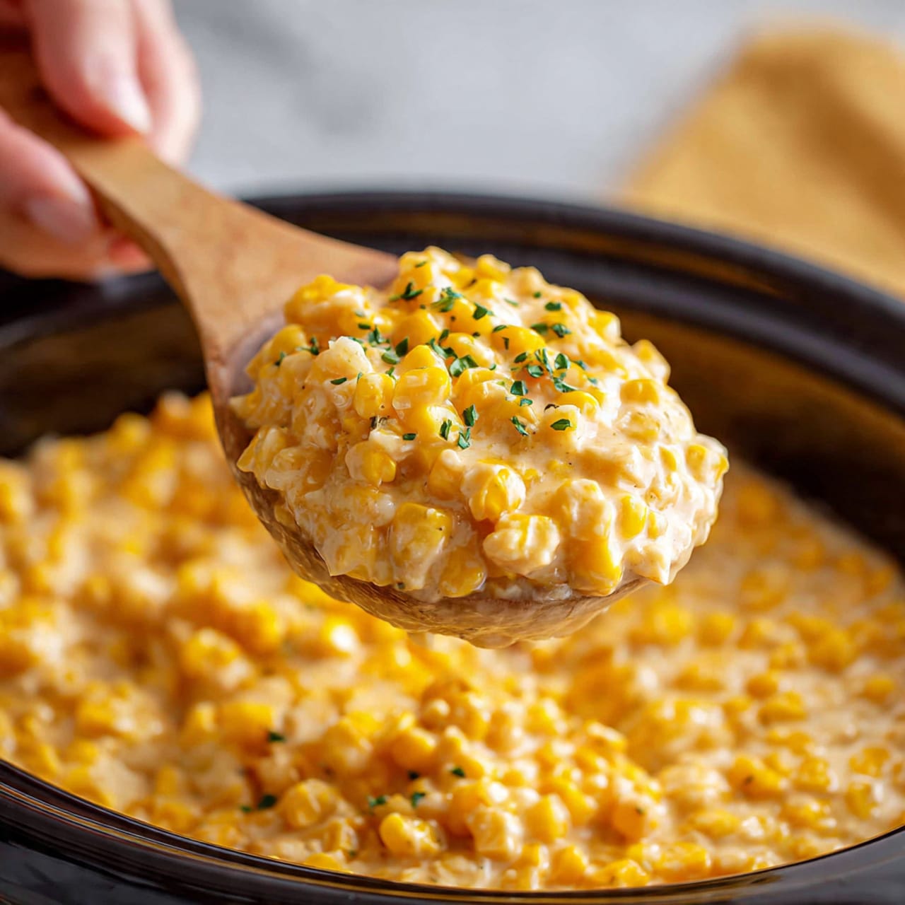 A close-up view of creamy slow cooker cheddar corn held by a woman's hand with a wooden spoon, showing a thick mixture of yellow corn kernels and melted light orange cheddar cheese, sprinkled with small bits of green herbs, all blended into a rich, slightly chunky texture. The background is blurred but has warm yellow and light brown colors with a white marbled texture beneath, emphasizing the cozy and comforting feel of the dish. photo taken with an iphone --ar 4:5 --v 7