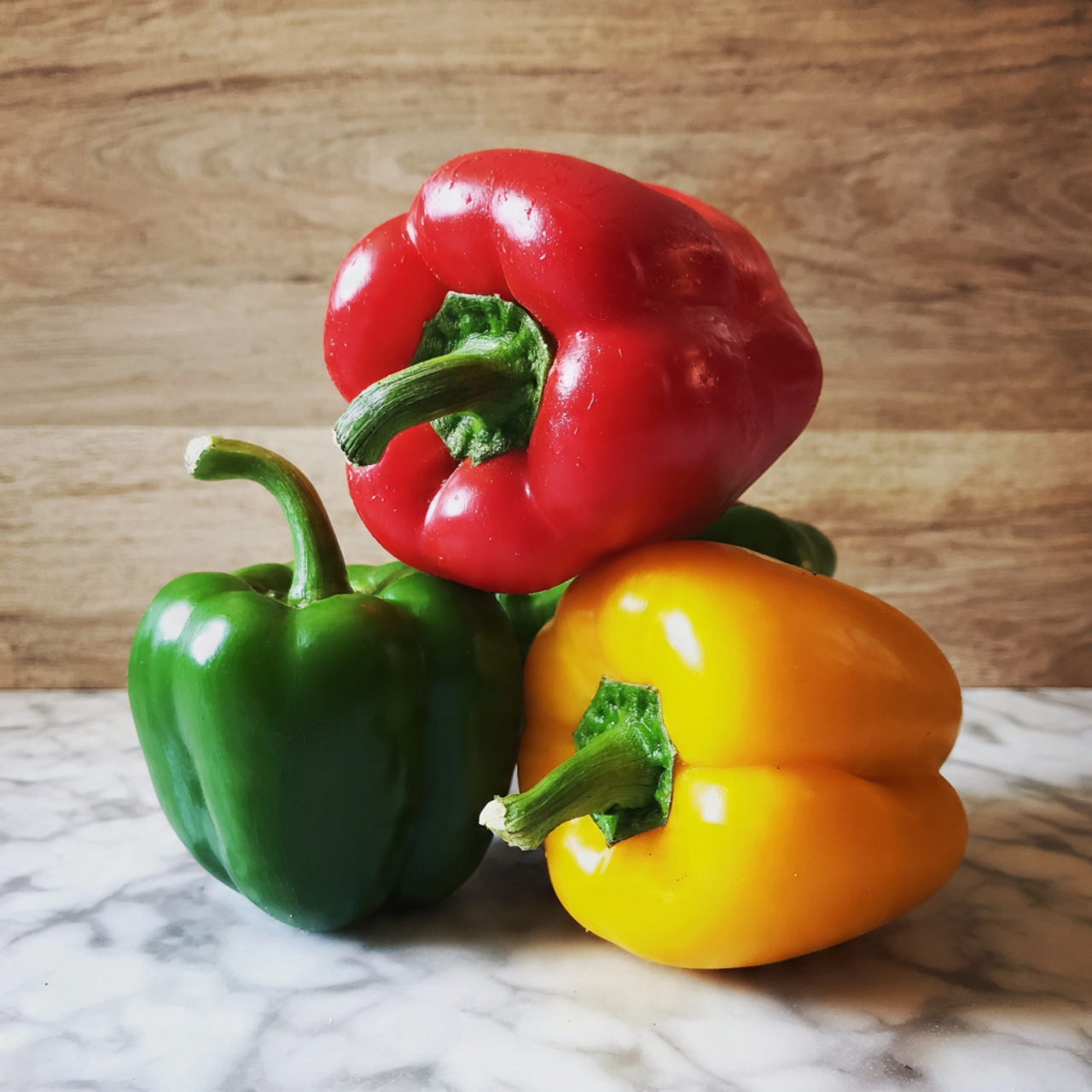 The image shows five bell peppers stacked together on a white marbled surface, with a wooden textured background. There are two large red bell peppers on top, with bright smooth skin and green stems. Below them, there is one green bell pepper on the left and one yellow bell pepper on the right, both with shiny surfaces and green stems. In front of these, slightly lower, there is another small green pepper, partially visible, with a shiny texture. Photo taken with an iphone --ar 4:5 --v 7
