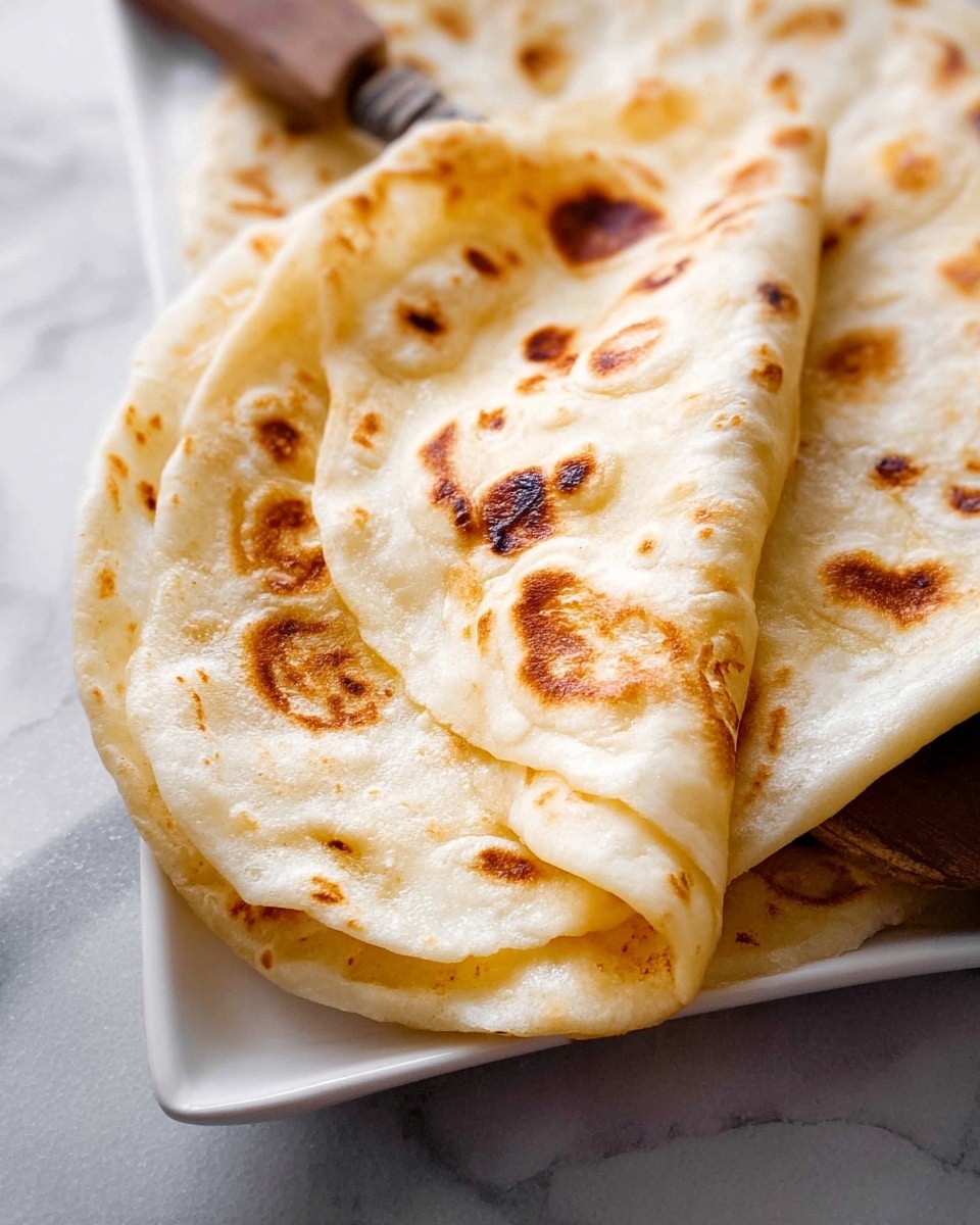 The image shows a close-up view of two soft flatbreads folded on a white rectangular plate. The flatbreads have a light golden color with darker brown char spots scattered across the surface, showing a slightly bubbly and fluffy texture. The top flatbread is lifted slightly at one edge, revealing a soft and slightly stretchy inside. The plate rests on a white marbled surface. Photo taken with an iphone --ar 4:5 --v 7
