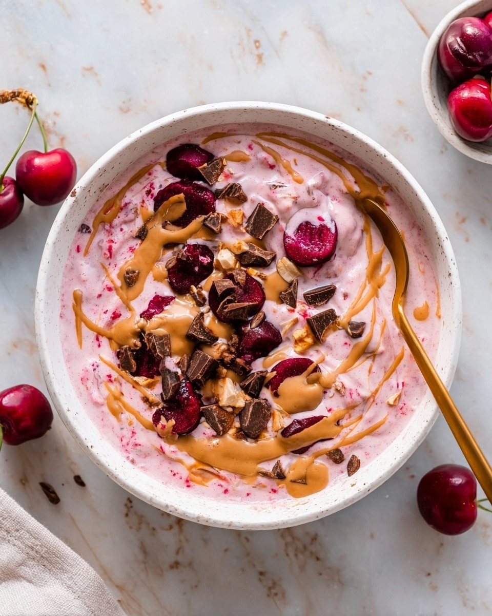 A white bowl with a creamy pink mix filling the base, made from a soft texture with visible white swirls and bits of dark red cherries mixed inside. On top, scattered small brown chocolate pieces and cut dark red cherries add a rich contrast. Light brown nut butter is drizzled over the whole dish in thin lines, enhancing the colors and textures. The bowl rests on a white marbled surface, with a golden spoon placed inside the bowl showing part of its handle. A few fresh dark red cherries are placed on the marble around the bowl, adding extra color and freshness to the scene photo taken with an iphone --ar 4:5 --v 7